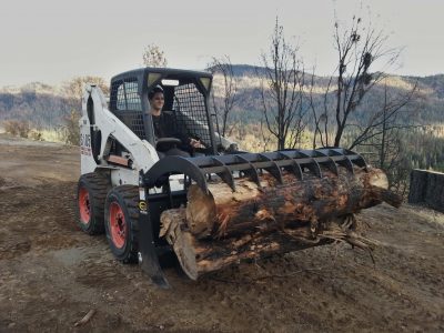 Image of a Bobcat skidsteer carrying several large logs with a grapple rake.