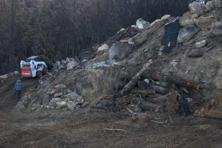 Image of the Bobcat driving up a steep road and a pile of logs in the foreground